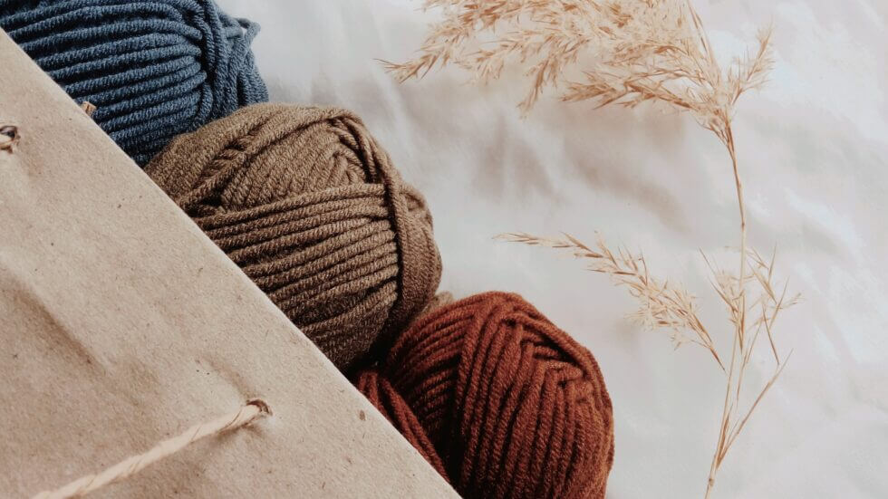 One skein each of blue, brown, and rust colored yarn peek out of a brown paper gift bag on top of white cloth.