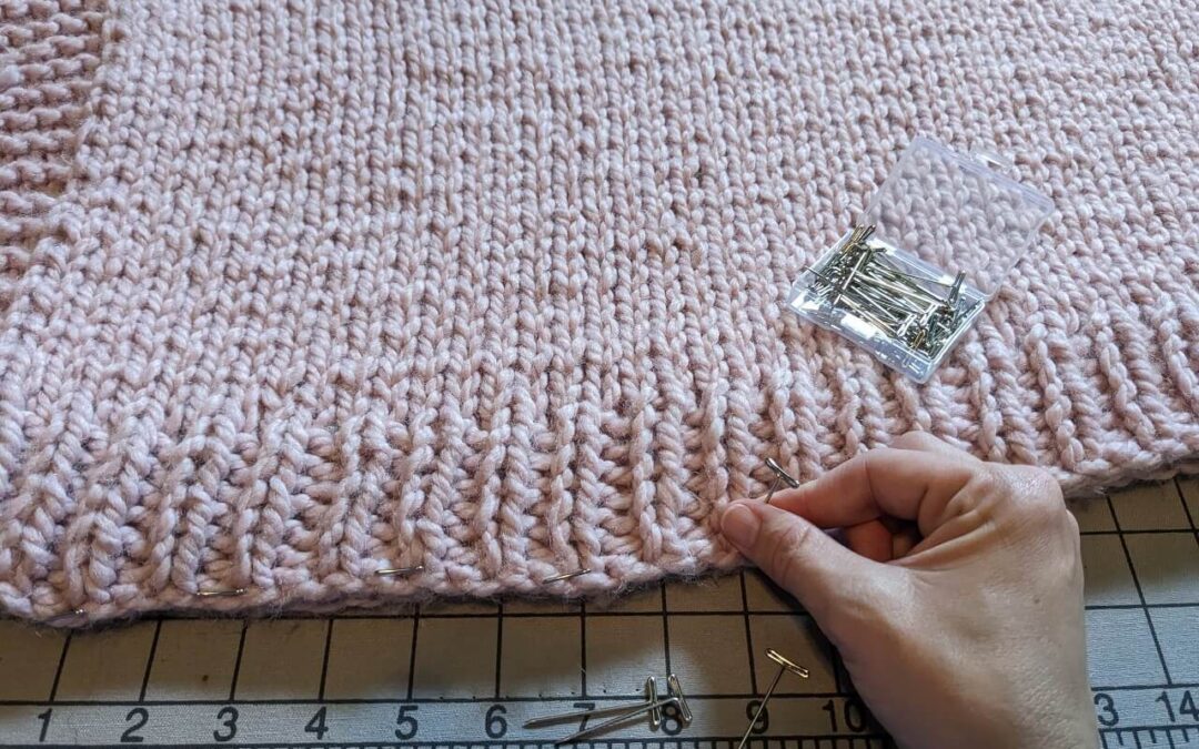 A hand is inserting T-pins through a pink knitted garment onto a blocking board.