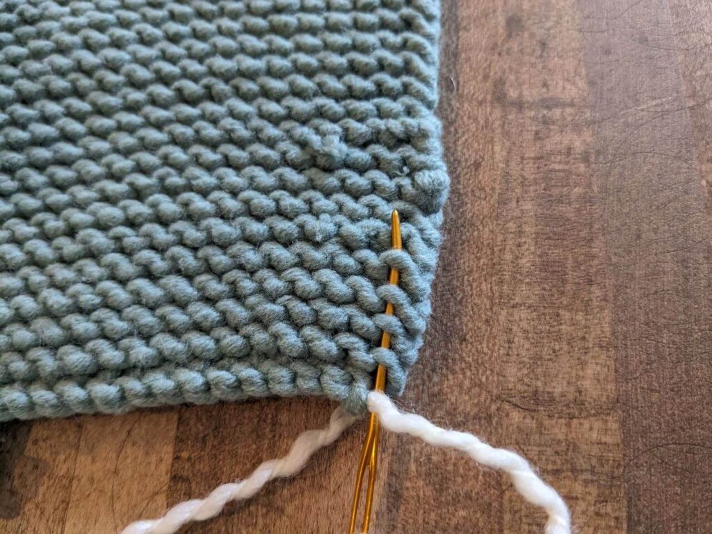 A gold yarn needle, threaded with cream yarn, is inserted through a couple of stitches of a teal garter stitch knitted piece.