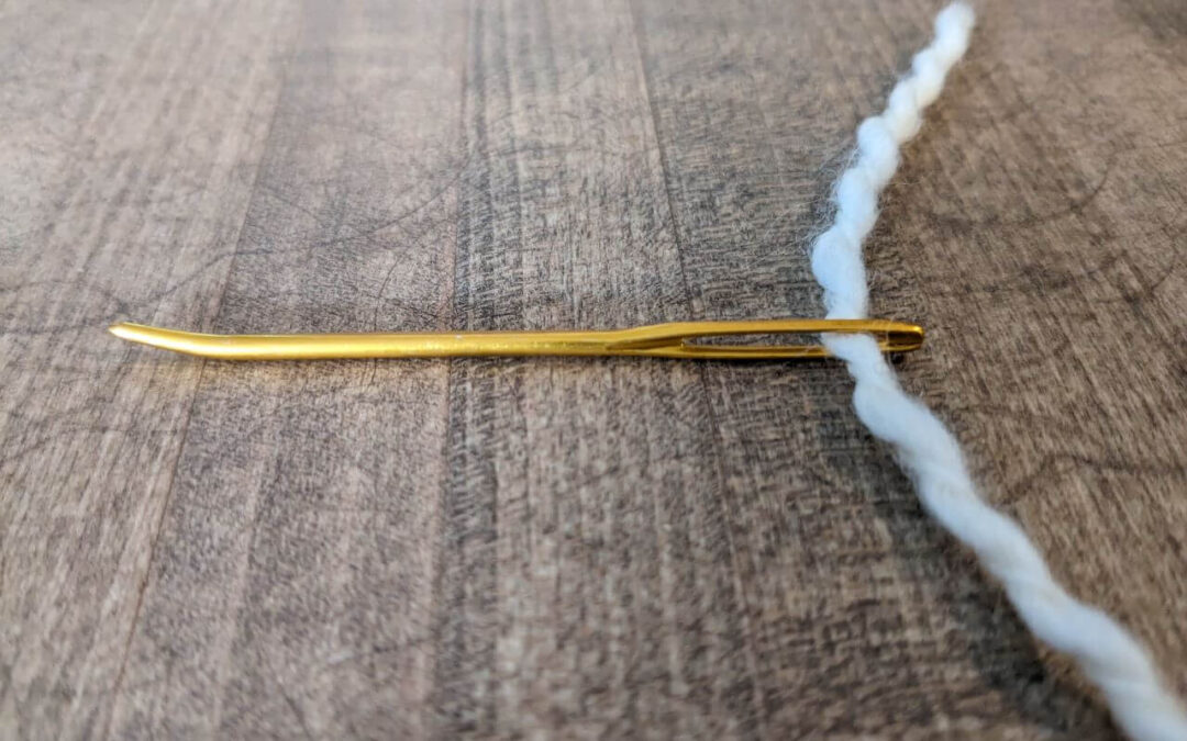 A gold yarn needle sits on a wooden counter, threaded with thick cream yarn, ready to start weaving.