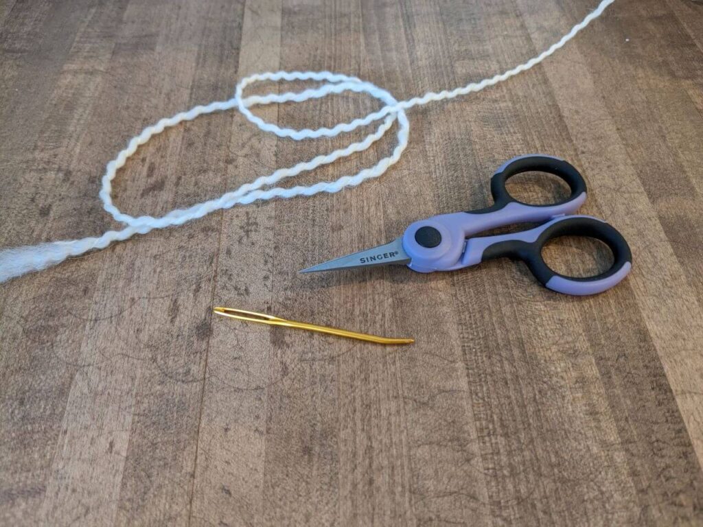 A gold yarn needle, small purple thread scissors, and a length of cream yarn sit next to each other on a wooden counter top.
